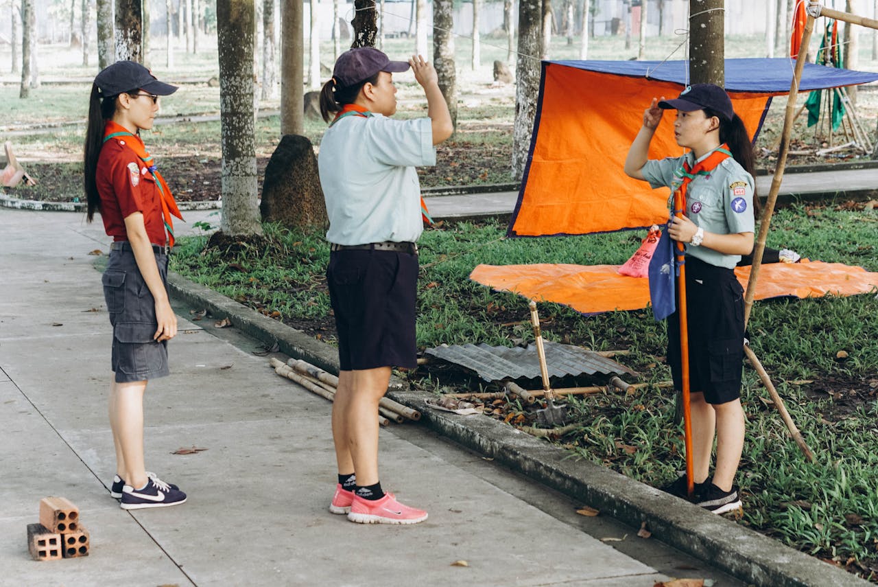 services-04 Three scouts salute in a park, participating in an outdoor scouting activity with orange tarp setup.